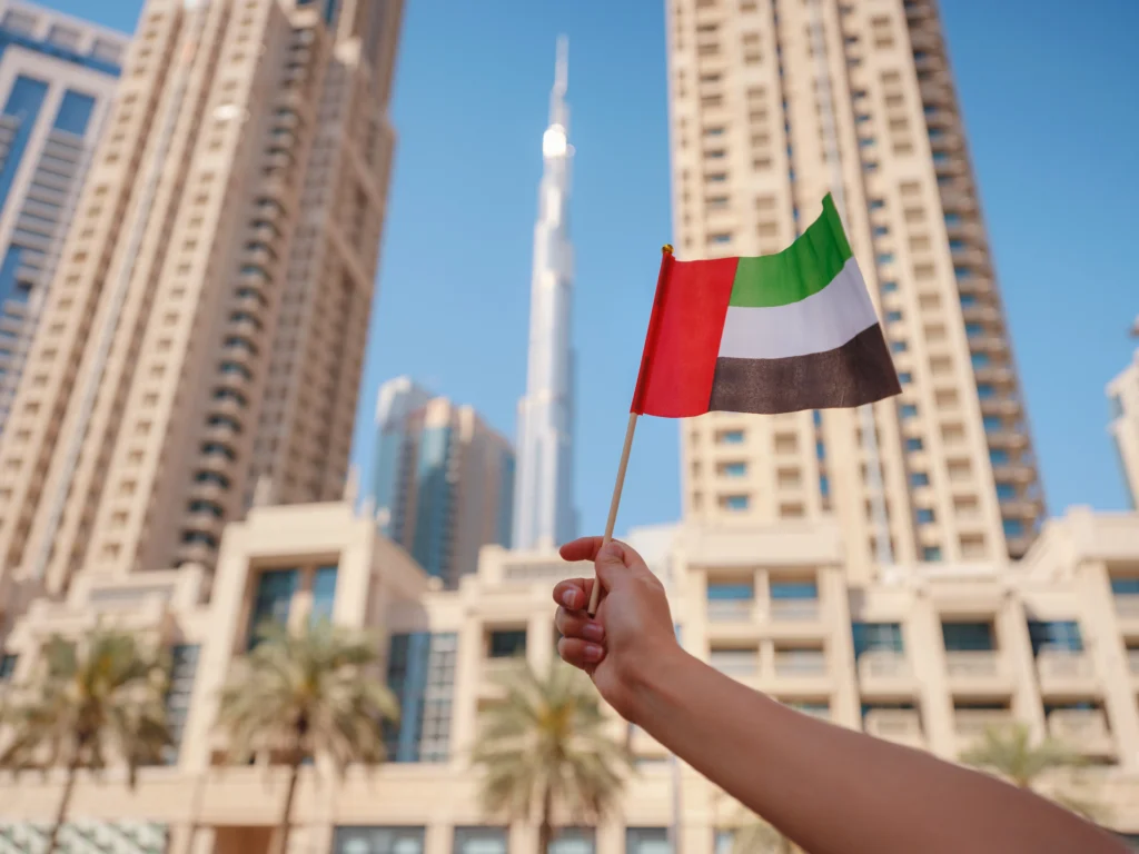 Hand holding a small UAE flag with the Burj Khalifa and downtown Dubai buildings in the background